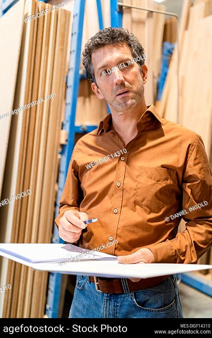Portrait of carpenter standing with ring binder in front of shelves with wooden planks