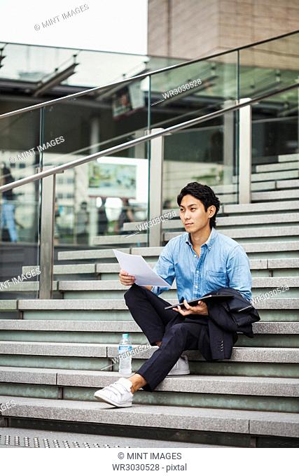 Businessman wearing blue shirt sitting outdoors on steps, holding digital tablet and papers