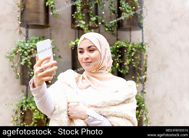 Young woman taking selfie through mobile phone in front of wall