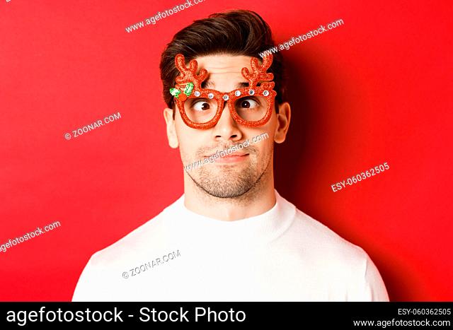 Concept of winter holidays, christmas and celebration. Close-up of funny brunette in party glasses, squinting and making faces, standing over red background