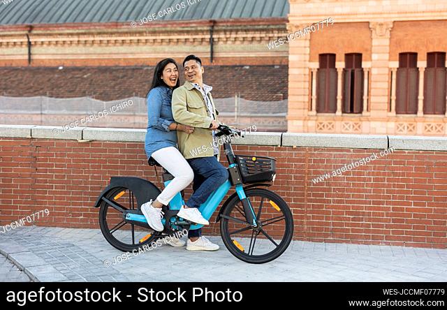 Happy mature couple sitting on electric bicycle by brick wall