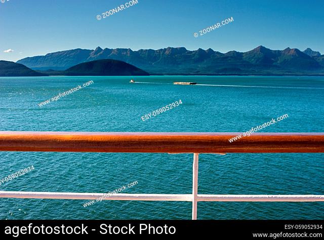 Tugboat pulling barge on a sunny day in Gastineau Channel, near Juneau, Alaska, USA. Douglas Island in background, cruise ship white metal and wooden railing in...