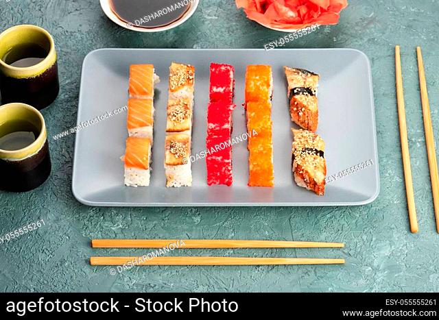 Sushi rolls and sashimi in a grey plate. Traditional Japanese cuisine with salmon, fish, wasabi, soy sauce, and ginger. Chopsticks and two cups of traditional...