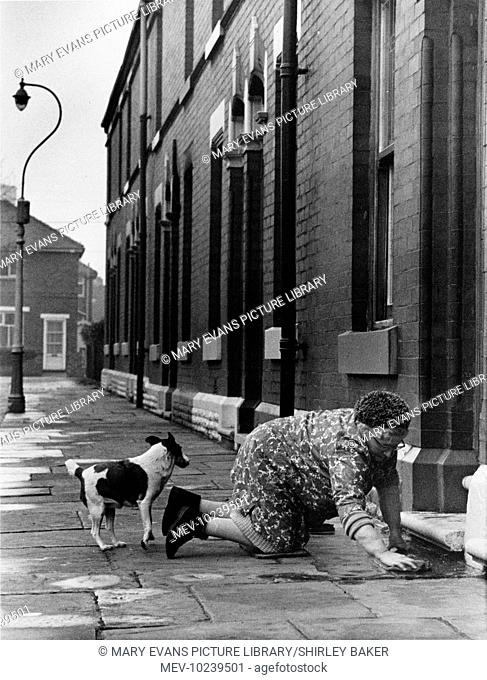 An elderly housewife in floral overall, fake fur hat and carpet slippers, kneels on the pavement in front of her terraced house in Manchester to clean the front...