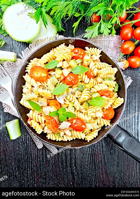 Fusilli with chicken, zucchini and tomatoes in a frying pan on burlap, forks, basil and parsley on wooden board background from above