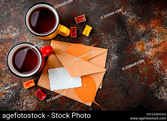 Two colored enamel cups of tea, two envelops, watercolors in cuvettes, , autumn leaves and bumps on a rusty brown background