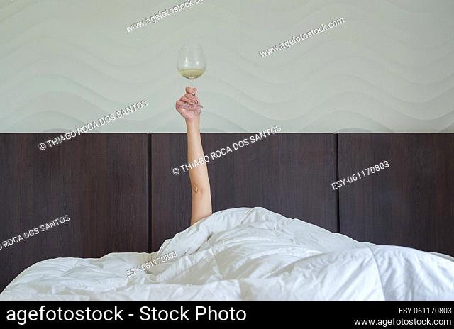Drinking wine early tomorrow. Woman in bed, under duvet, with arm raised up holding glass of wine. Morning wine in bed