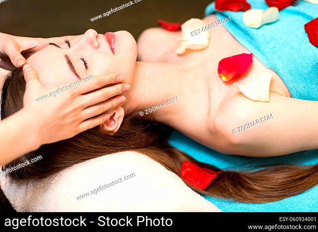 Young beautiful woman receiving head face massage in beauty spa with rose flower petals
