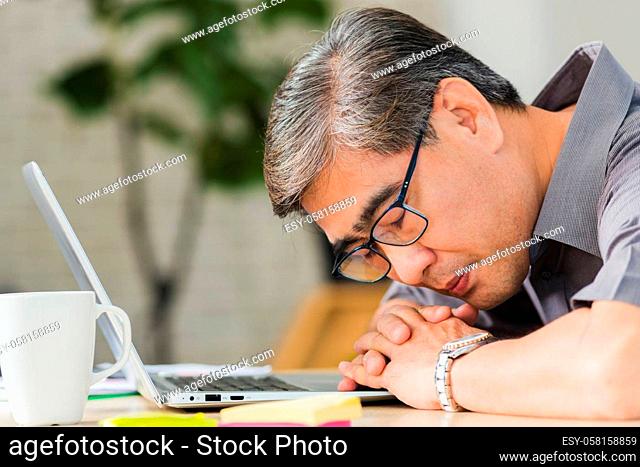 Asian businessman tired overworked he sleeping over a laptop computer on the desk. senior man with eyeglasses lying asleep on table at his working place