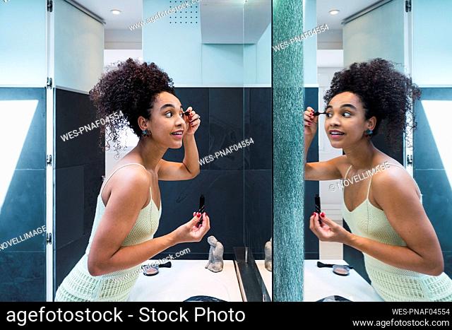 Smiling woman applying mascara standing in front of mirror at home