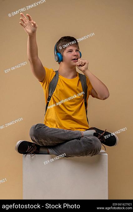 Focused cute teenage boy seated cross-legged on the box performing his favorite song to music