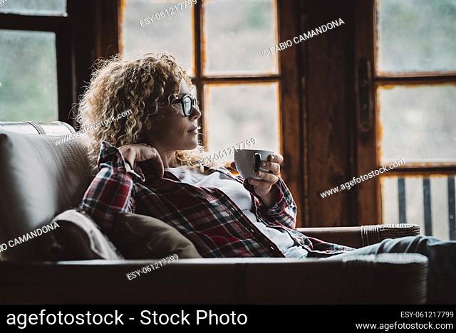 Pretty adult hipster woman sitting and relaxing at home with cup of tea and windows in background - relaxed and enjoyed female people in cozy cabin house for...