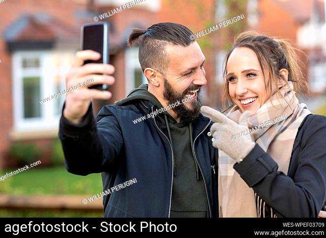 Young woman gesturing while taking selfie with cheerful boyfriend outside new house