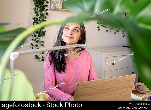 Smiling woman day dreaming while sitting with laptop at home
