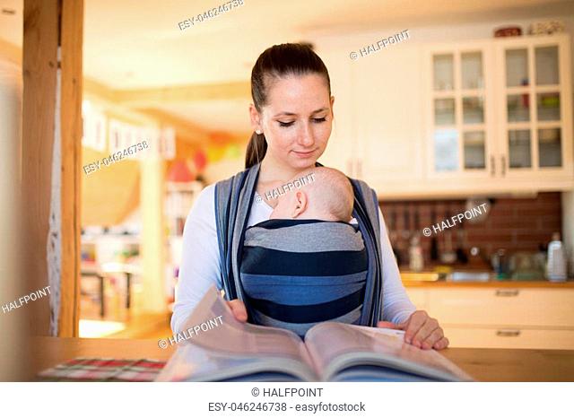Beautiful young mother in kitchen with her baby son sleeping in sling at home, reading a book or studying something