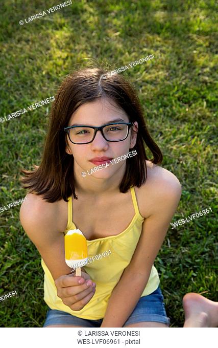 Portrait of girl wearing glasses sitting on a meadow eating ice lolly