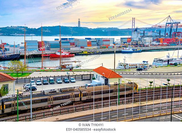 Train on railroad by comercial port with ships and containers, 25 de Abril Bridge over Tagus river at sunset, Christ the King monument on background, Lisbon