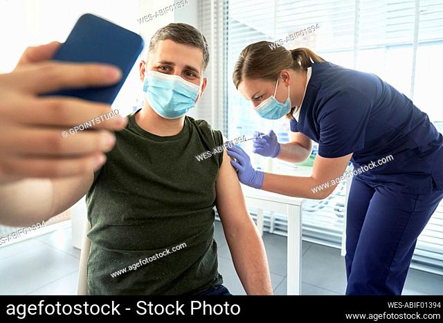 Man with face mask taking selfie through smart phone during COVID-19 vaccination