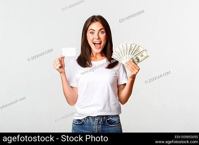 Excited happy brunette girl showing credit card and money, smiling over white background
