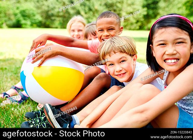 Multikulturelle Gruppe glücklicher Kinder sitzt mit Ball im Sommer auf einer Wiese