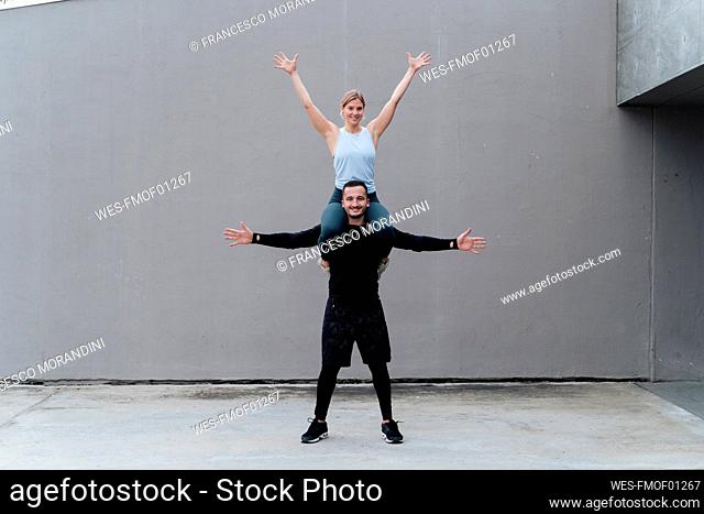 Woman with hand raised sitting on man shoulder standing with arms outstretched against wall
