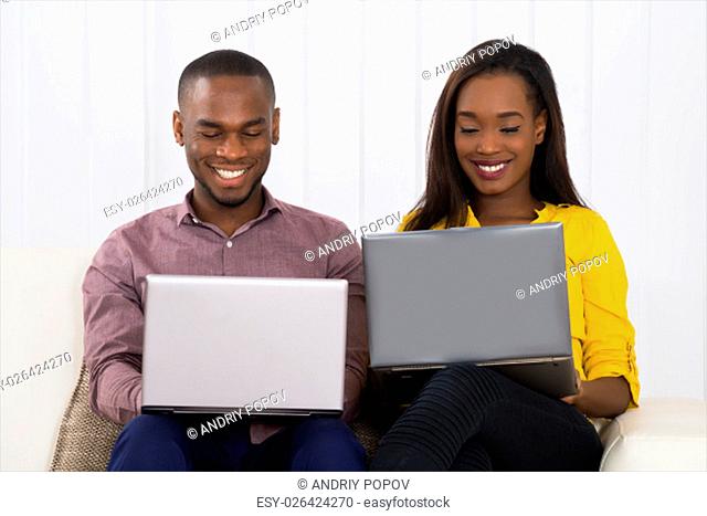 Happy Young Couple Using Laptops While Sitting On Sofa