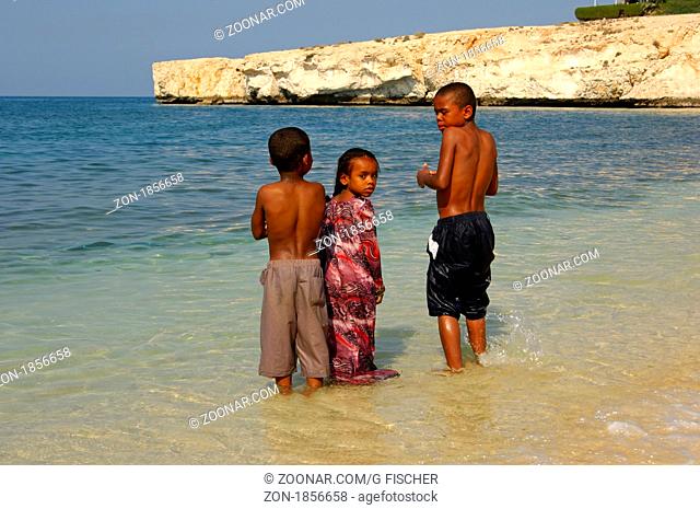 Zwei einheimische Jungen und ein Mädchen stehen am Ufer im Wasser, das Mädchen blickt neugierig zur Kamera, Maskat, Golf von Oman