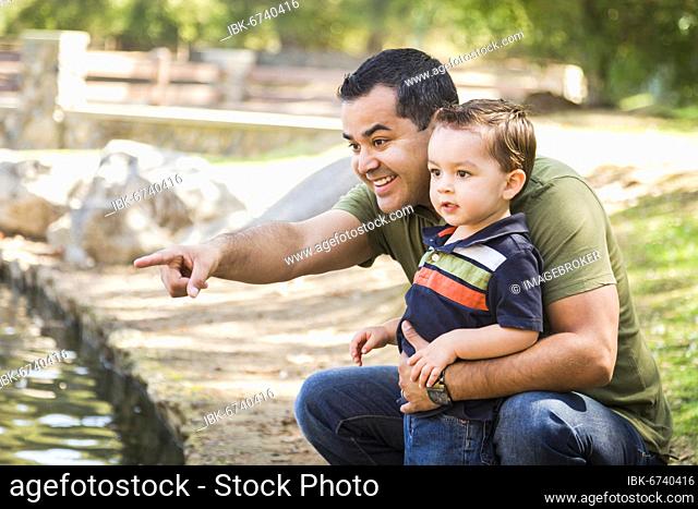 Happy hispanic father points with mixed-race son at the park pond