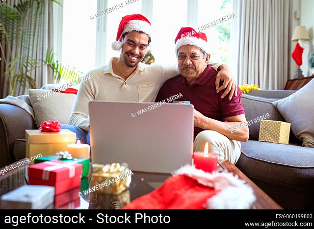 Happy biracial adult son and senior father in santa hats making laptop christmas video call
