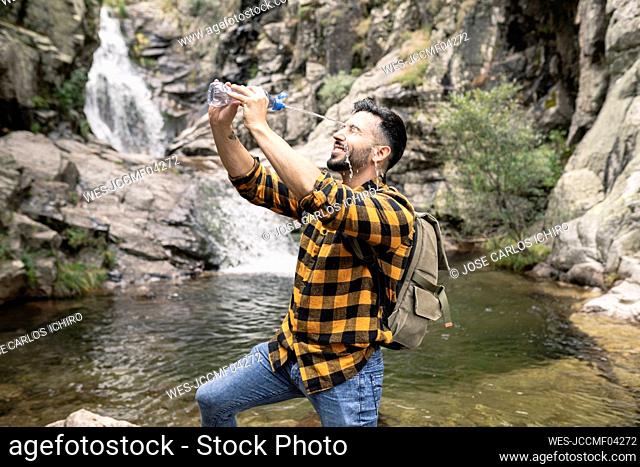 Smiling male tourist putting water on face through bottle by pond