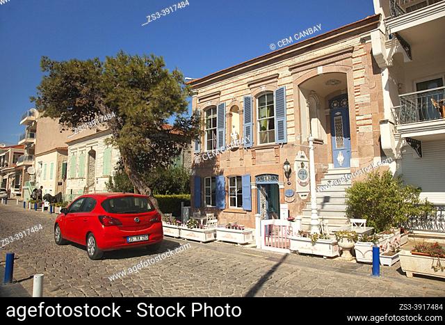 Car in front of the traditional houses on Reha Midilli Street at Smaller Sea-Küçükdeniz district of Old Foca with a woman on a bike in the foreground
