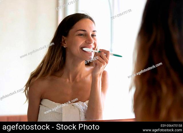 Smiling young woman in towel after shower look in mirror brush clean teeth with toothbrush in bright home bath, happy millennial girl do facial daily morning...