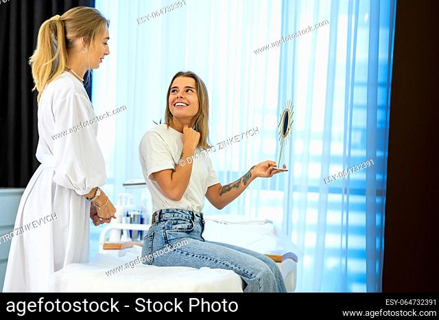 Smiling female client satisfied with result after cosmetic procedures sitting on couch near doctor holding mirror