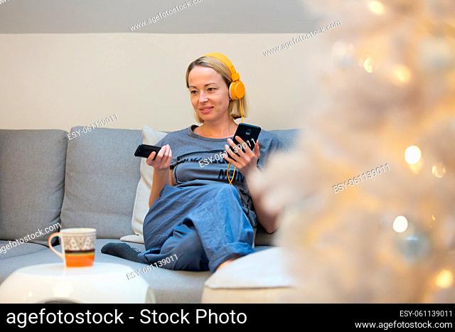 Young cheerful woman sitting indoors at home living room sofa watching TV, using social media on phone for video chatting and staying connected with her loved...