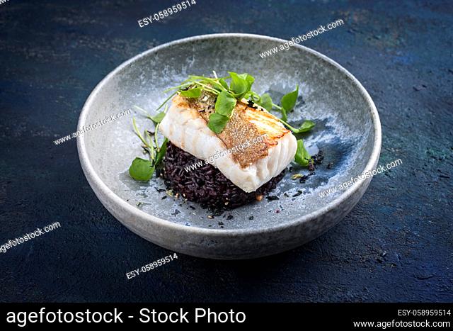 Modern style traditional fried skrei cod fish filet with portulaca lettuce, and black rice served as close-up on ceramic design plate with copy space
