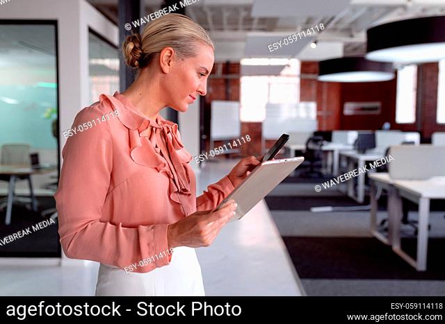 Happy caucasian businesswoman standing in office holding tablet and using smartphone