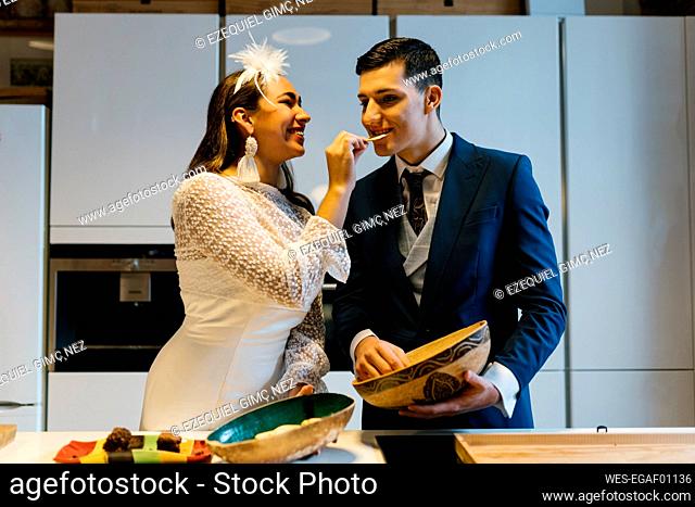 Smiling bride feeding potato chip to groom while standing in kitchen at home