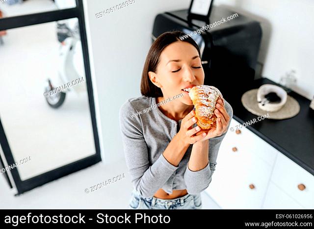 Portrait of rejoicing woman eats tasty croissant at home. Unhealthy food concept