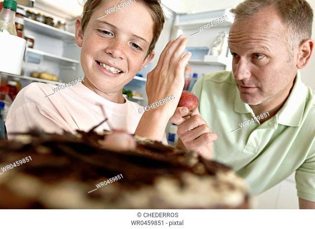 Boy standing in front of a cake and refusing an apple from his father