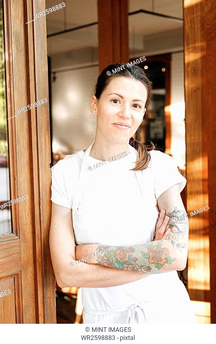 Portrait of a woman wearing a white apron standing with her arms crossed in a doorway, tattoos on her left arm