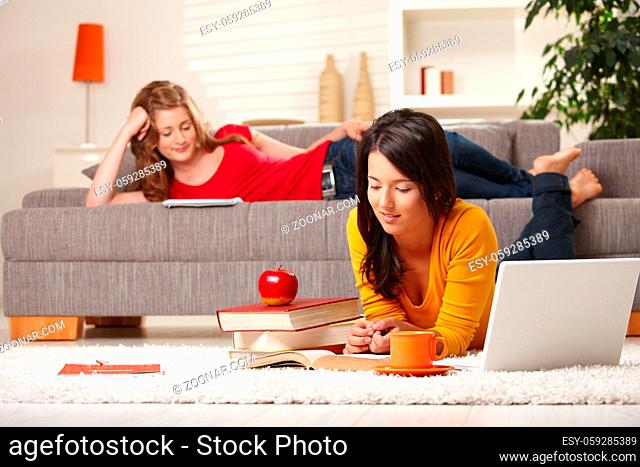 Teenage girls studying at home in living room lying on sofa and floor with books and laptop