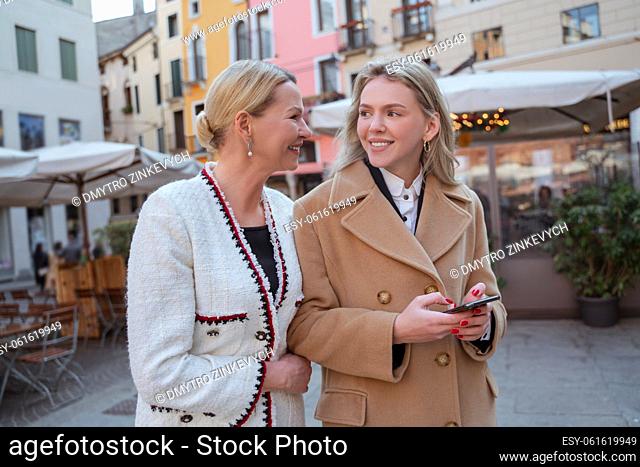 Smiling beautiful blonde woman with the smartphone standing beside a stylish mature lady in the pedestrian zone