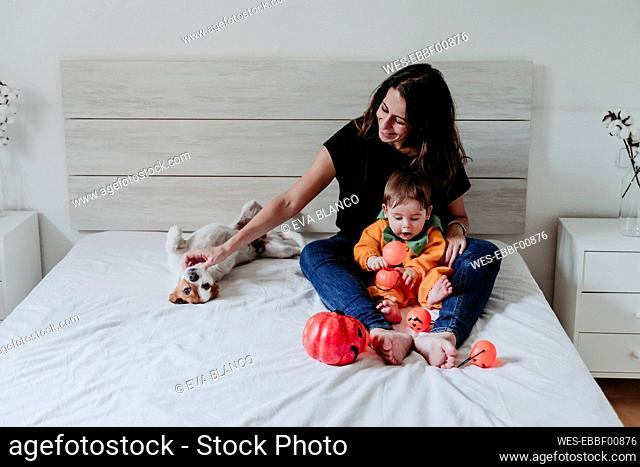 Mother sitting with son weraing halloween costume and dog lying on bed at home