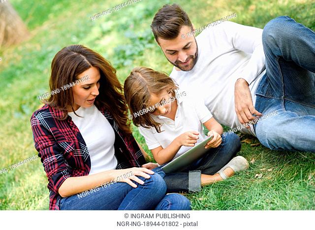 Happy family in a urban park playing with tablet computer. Father, mother and little daughter sitting on the grass laughing