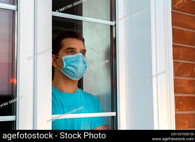 Young man in medical mask is looking out window. Coronavirus pandemic. Home quarantine, self-isolation because of the Coronavirus disease, COVID-19