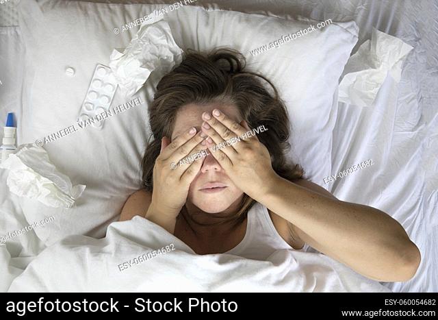 A sick young woman in bed, lying with her head under the blankets and a pile of tissues next to her top view. Fever staying home from work