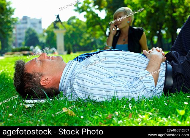 Businessman resting in grass in park, busineswoman talking on mobile in the background
