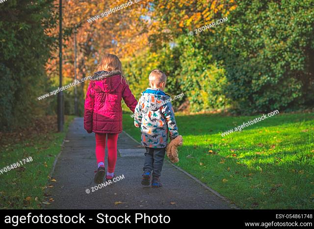Boy holding hs favourite teddy bear soft toy holding hands with his sister and walking together on a path in the countryside in autumn