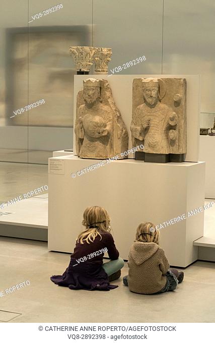 Two light haired children sat cross legged in front of two light stone crowned figure carvings, listening to historical commentaries, Louvre-Lens Museum