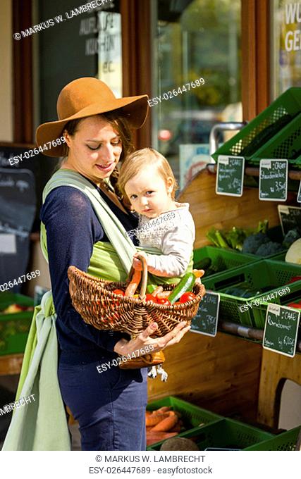 A young mother carrying her little baby in a sling before belly and buys vegetables at a market stall of a Health Food Shop